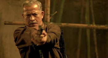 Movie still from “Ong-Bak: The Thai Warrior” (2003), directed by Prachya Pinkaew – An older man is holding a gun and aiming at the camera; Close Up shot, Low angle