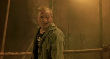 Movie still from “Ong-Bak: The Thai Warrior” (2003), directed by Prachya Pinkaew – A man in a green shirt is standing in front of a wall; Medium shot, Over the shoulder angle