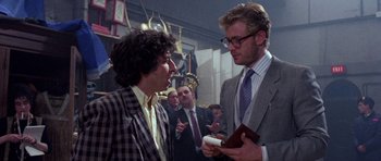 Movie still from “Opera” (1987), directed by Dario Argento – Two men in suits talking to each other in front of a group of people; Medium shot, Over the shoulder angle