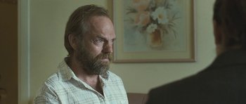 Movie still from “Oranges and Sunshine” (2010), directed by Jim Loach – A man with a beard is sitting in front of a painting; Close Up shot, Over the shoulder angle