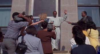 Movie still from “Original Gangstas” (1996), directed by Larry Cohen – A group of people gathered around a man holding a microphone; Wide shot, High angle