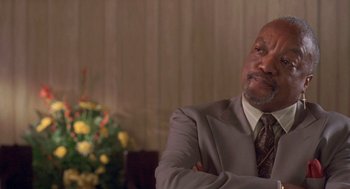 Movie still from “Original Gangstas” (1996), directed by Larry Cohen – A man in a suit and tie sitting in front of a flower arrangement; Close Up shot, Low angle