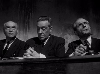 Movie still from “Orpheus” (1950), directed by Jean Cocteau – A group of men sitting next to each other in front of a wall; Medium shot, Low angle
