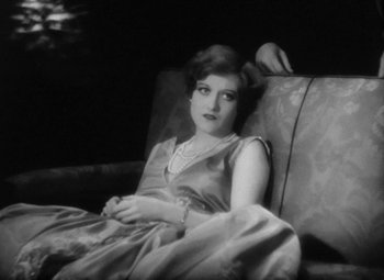 Movie still from “Our Dancing Daughters” (1928), directed by Harry Beaumont – A woman sitting on top of a couch in front of a window; Medium shot, High angle