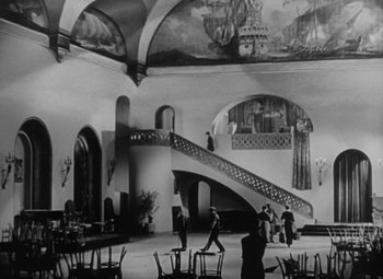 Movie still from “Our Dancing Daughters” (1928), directed by Harry Beaumont – A black - and - white photo of a room with chairs , tables , and a staircase; Extreme Wide shot, Low angle