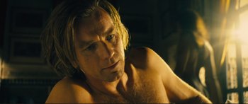 Movie still from “Our Kind of Traitor” (2016), directed by Susanna White – A man with long blonde hair and no shirt is posing for the camera; Close Up shot, Over the shoulder angle