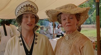 Movie still from “Out of Africa” (1985), directed by Sydney Pollack – Two women dressed in old fashion clothing standing next to each other in front of an audience; Close Up shot, Low angle