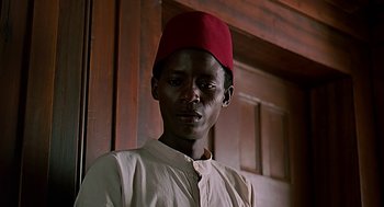 Movie still from “Out of Africa” (1985), directed by Sydney Pollack – A man wearing a red hat standing in front of a door; Close Up shot, Low angle