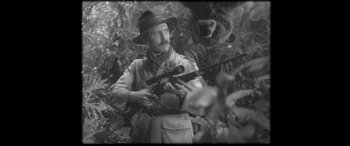 Movie still from “Paddington” (2014), directed by Paul King – A man holding a rifle in his hands while standing next to a brown bear; Medium shot, Low angle