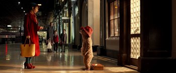 Movie still from “Paddington” (2014), directed by Paul King – A teddy bear wearing a red hat standing in front of a suitcase; Wide shot, Over the shoulder angle