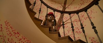 Movie still from “Paddington” (2014), directed by Paul King – A teddy bear wearing a red hat sitting on top of a spiral stair case railing; Wide shot, Low angle