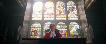 Movie still from “Paddington” (2014), directed by Paul King – A judge sitting in front of a stained glass window; Medium shot, Low angle