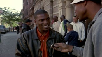 Movie still from “Paid in Full” (2002), directed by Charles Stone III – A man smiles while holding a cell phone in front of a crowd; Medium shot, Over the shoulder angle