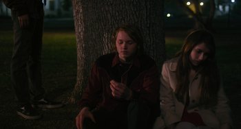 Movie still from “Palo Alto” (2013), directed by Gia Coppola – A man sitting next to a tree at night; Medium shot, Over the shoulder angle