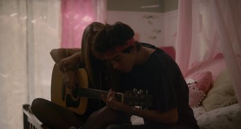 Movie still from “Palo Alto” (2013), directed by Gia Coppola – A man playing a guitar while a woman sits next to him on a couch; Medium shot, Over the shoulder angle