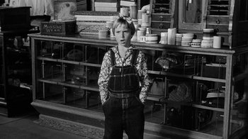 Movie still from “Paper Moon” (1973), directed by Peter Bogdanovich – A young boy in overalls standing in front of shelves; Medium shot, Over the shoulder angle