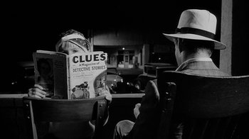 Movie still from “Paper Moon” (1973), directed by Peter Bogdanovich – A man sitting in front of another man reading a magazine; Medium shot, High angle