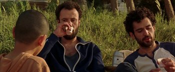 Movie still from “Paradise Now” (2005), directed by Hany Abu-Assad – A man sitting on the ground drinking from a cup; Medium shot, Low angle