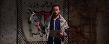 Movie still from “Paradise Now” (2005), directed by Hany Abu-Assad – A man holding a gun and a book in front of an arabic flag; Medium shot, Low angle