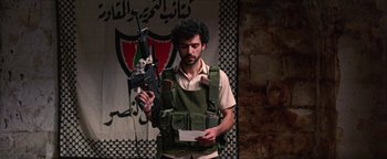 Movie still from “Paradise Now” (2005), directed by Hany Abu-Assad – A man holding an ar - 1 5 rifle while standing in front of a flag; Medium shot, Over the shoulder angle