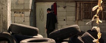 Movie still from “Paradise Now” (2005), directed by Hany Abu-Assad – A man standing in front of a door next to a pile of tires; Wide shot, Over the shoulder angle