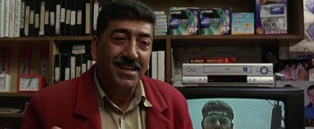 Movie still from “Paradise Now” (2005), directed by Hany Abu-Assad – A man in a red jacket is sitting in front of a dvd shelf; Close Up shot, Over the shoulder angle