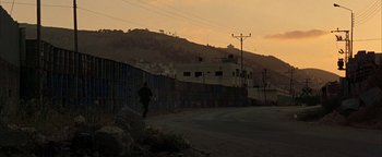 Movie still from “Paradise Now” (2005), directed by Hany Abu-Assad – A person walking on the side of a road near a wall; Extreme Wide shot, Low angle