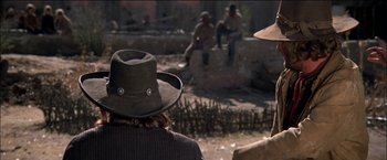 Movie still from “Pat Garrett & Billy the Kid” (1973), directed by Sam Peckinpah – A man wearing a cowboy hat sitting on a stone wall; Medium shot, Over the shoulder angle