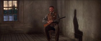 Movie still from “Pat Garrett & Billy the Kid” (1973), directed by Sam Peckinpah – A man sitting on a chair holding a rifle; Medium shot, Low angle