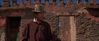 Movie still from “Pat Garrett & Billy the Kid” (1973), directed by Sam Peckinpah – A man wearing a hat standing in front of a brick building; Medium shot, Low angle
