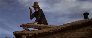 Movie still from “Pat Garrett & Billy the Kid” (1973), directed by Sam Peckinpah – A man in a cowboy hat holding a gun and standing on top of a wooden fence; Medium shot, Low angle