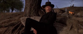 Movie still from “Pat Garrett & Billy the Kid” (1973), directed by Sam Peckinpah – A man sitting on a rock in front of a tree; Medium shot, Low angle