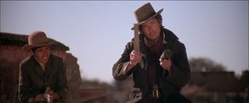 Movie still from “Pat Garrett & Billy the Kid” (1973), directed by Sam Peckinpah – A man in a cowboy hat holding an axe and a microphone; Medium shot, Low angle