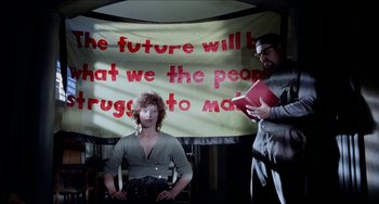 Movie still from “Patty Hearst” (1988), directed by Paul Schrader – Two people in front of a banner that says " the future will be what we the people struggle to make "; Medium shot, Low angle
