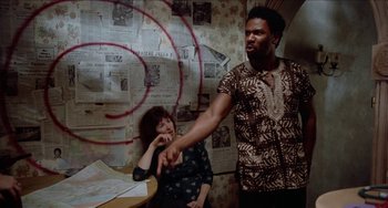 Movie still from “Patty Hearst” (1988), directed by Paul Schrader – A man and a woman pointing at each other in front of a wall covered in newspaper; Medium shot, High angle