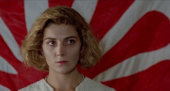 Movie still from “Patty Hearst” (1988), directed by Paul Schrader – A young woman with short brown hair wearing a white shirt and standing in front of a red and white flag; Close Up shot, Low angle