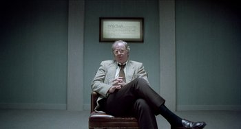 Movie still from “Patty Hearst” (1988), directed by Paul Schrader – An older man sitting in a chair in front of a wall; Medium shot, Over the shoulder angle