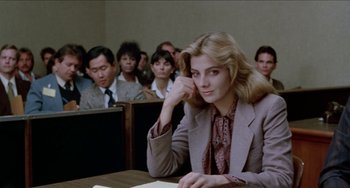 Movie still from “Patty Hearst” (1988), directed by Paul Schrader – A woman sitting at a table in front of a group of people; Medium shot, Over the shoulder angle