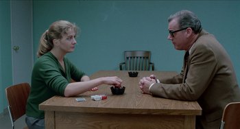 Movie still from “Patty Hearst” (1988), directed by Paul Schrader – Two people sitting at a wooden table playing cards; Medium shot, Over the shoulder angle