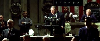Movie still from “Pearl Harbor” (2001), directed by Michael Bay – A man in a suit and tie speaking at a podium with microphones in front of him; Medium shot, Over the shoulder angle