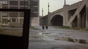 Movie still from “Pennies from Heaven” (1981), directed by Herbert Ross – Two people riding motorcycles down a street near a bridge; Extreme Wide shot, Over the shoulder angle