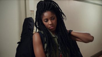 Movie still from “People Places Things” (2015), directed by Jim Strouse – A woman with long dreadlocks holding a guitar case; Close Up shot, Over the shoulder angle