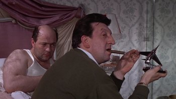 Movie still from “Performance” (1970), directed by Donald Cammell – A man smoking a cigar while another man watches; Medium shot, Over the shoulder angle
