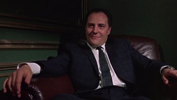 Movie still from “Performance” (1970), directed by Donald Cammell – A man in a suit and tie sitting on a couch; Medium shot, Low angle