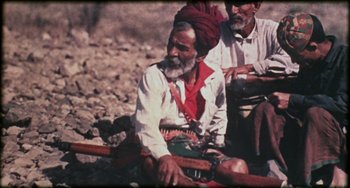 Movie still from “Performance” (1970), directed by Donald Cammell – A man sitting on the ground while holding a rifle; Medium shot, High angle