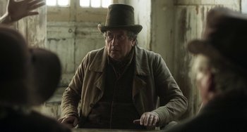 Movie still from “Peterloo” (2018), directed by Mike Leigh – An older man wearing a top hat sitting at a table; Medium shot, Over the shoulder angle