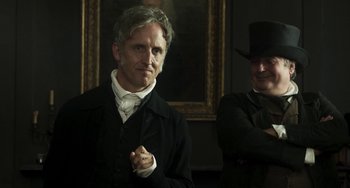 Movie still from “Peterloo” (2018), directed by Mike Leigh – A man standing next to another man wearing a top hat; Medium shot, Over the shoulder angle
