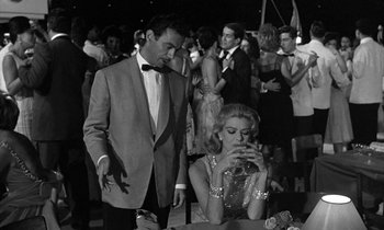 Movie still from “Phaedra” (1962), directed by Jules Dassin – A man and a woman in formal wear standing in front of a crowd; Medium shot, Low angle