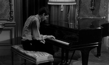 Movie still from “Phaedra” (1962), directed by Jules Dassin – A man sitting at a grand piano in front of a lamp; Medium shot, High angle