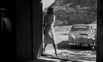 Movie still from “Phaedra” (1962), directed by Jules Dassin – A black and white photo of a woman standing in front of an old car; Wide shot, Low angle